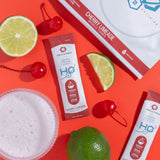 H2O Electrolytes™ Cherry Limeade zero sugar electrolyte powder with fresh cherries, lime slices, and a fizzy hydration drink on a striking red background. Sugar-free electrolyte mix for peak hydration and endurance.