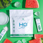 H2O Electrolytes™ Watermelon zero sugar electrolyte powder with fresh watermelon slices, ice cubes, and a fizzy mixed drink on a vibrant green background. Hydration packets for fast replenishment and optimal performance.