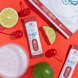 H2O Electrolytes™ Cherry Limeade zero sugar electrolyte powder with fresh cherries, lime slices, and a fizzy hydration drink on a striking red background. Sugar-free electrolyte mix for peak hydration and endurance.